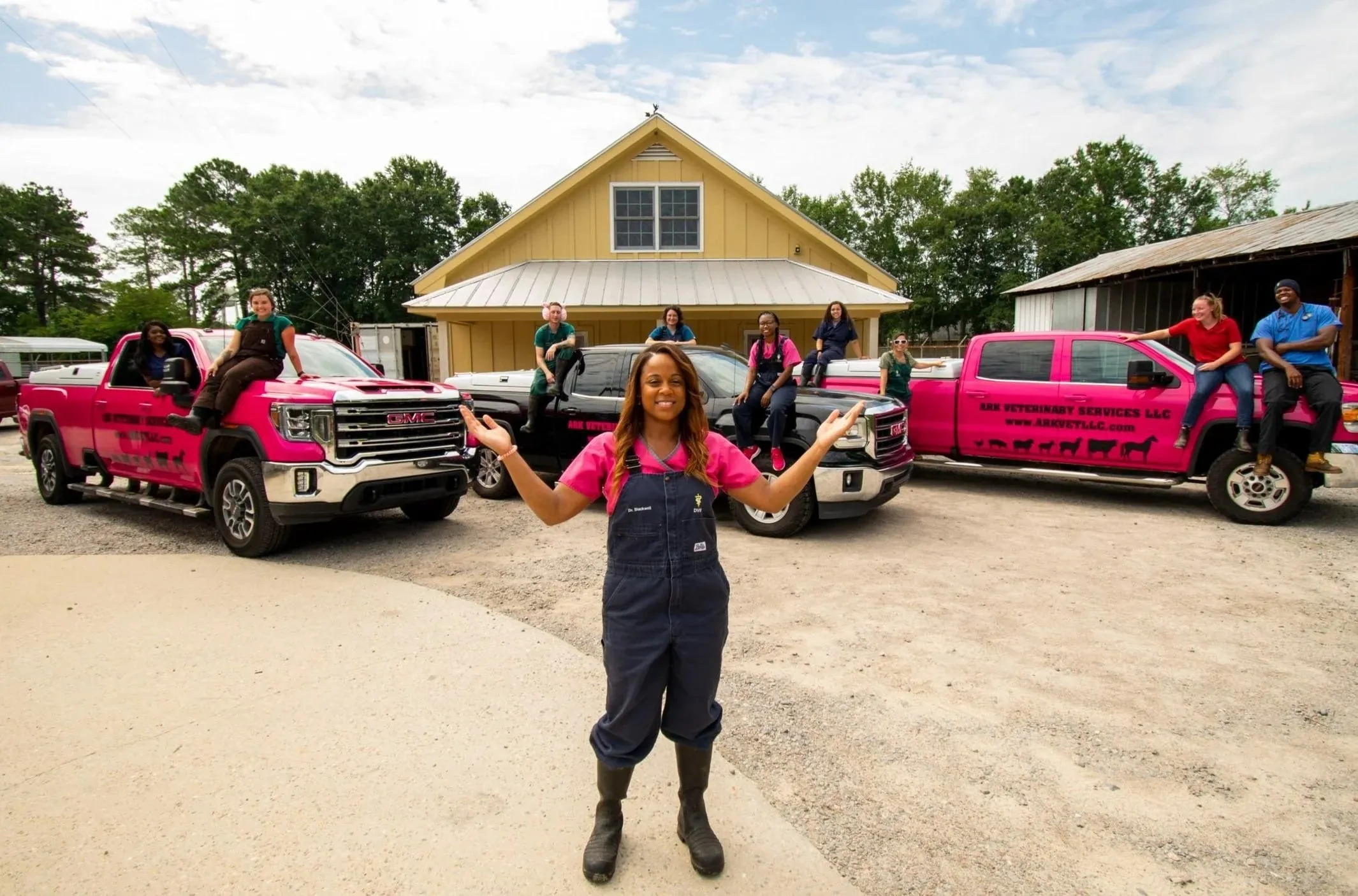 Dr. Sarah Blackwell in pink scrubs and overalls with arms open in front of the Ark team, who sit on three hot pink and black pickup trucks outside a yellow barn